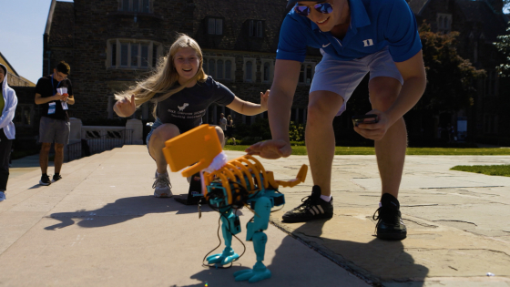 a small robotic orange and blue t-rex on cement stairs with people standing next to it