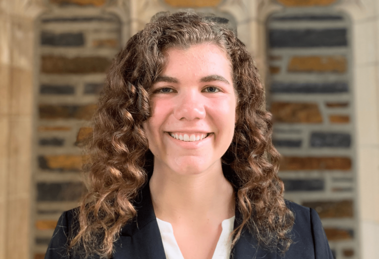 woman with long brown curly hair in dark jacket in front of Duke stone arch