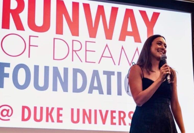 woman with long dark hair holding microphone on stage with Runway of Dream Foundation @ Duke University sign as backdrop