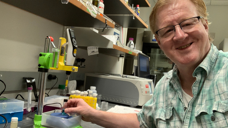A man smiling at the camera while working with vials in a wet lab