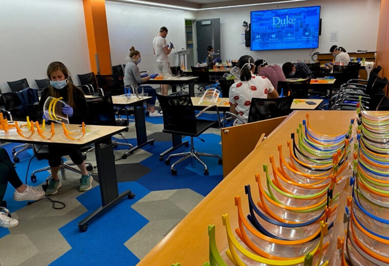 People crowd around desks working on face shields, which are piled up in a row in the corner