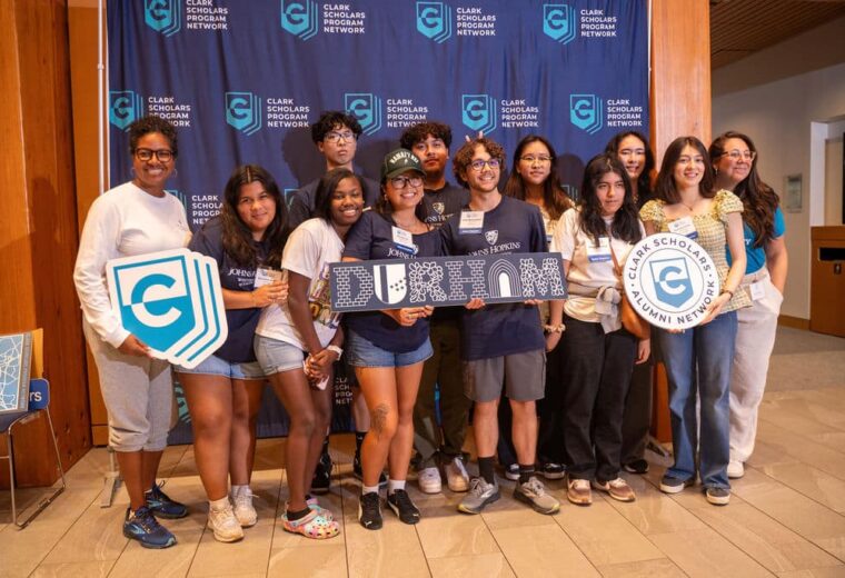 A group of twelve scholars smile in front of a blue banner that reads “Clark Scholars Program Network.” Several of them hold signs, including one with the Johns Hopkins University logo, another that says “Durham,” and a circular one that reads “Clark Scholars Alumni Network.”