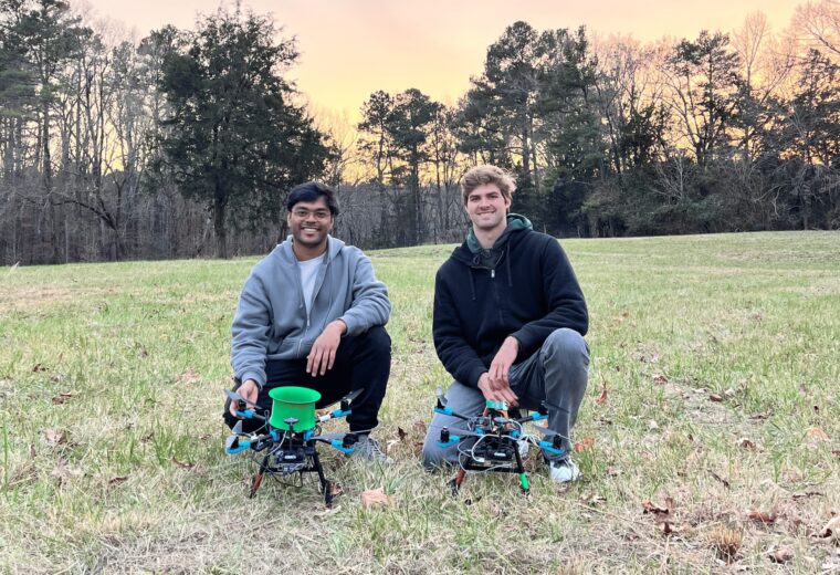 two people kneel in a grassy field with two flying drones
