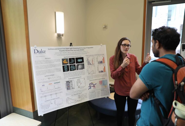 a person speaks while another listens, in front of a research poster
